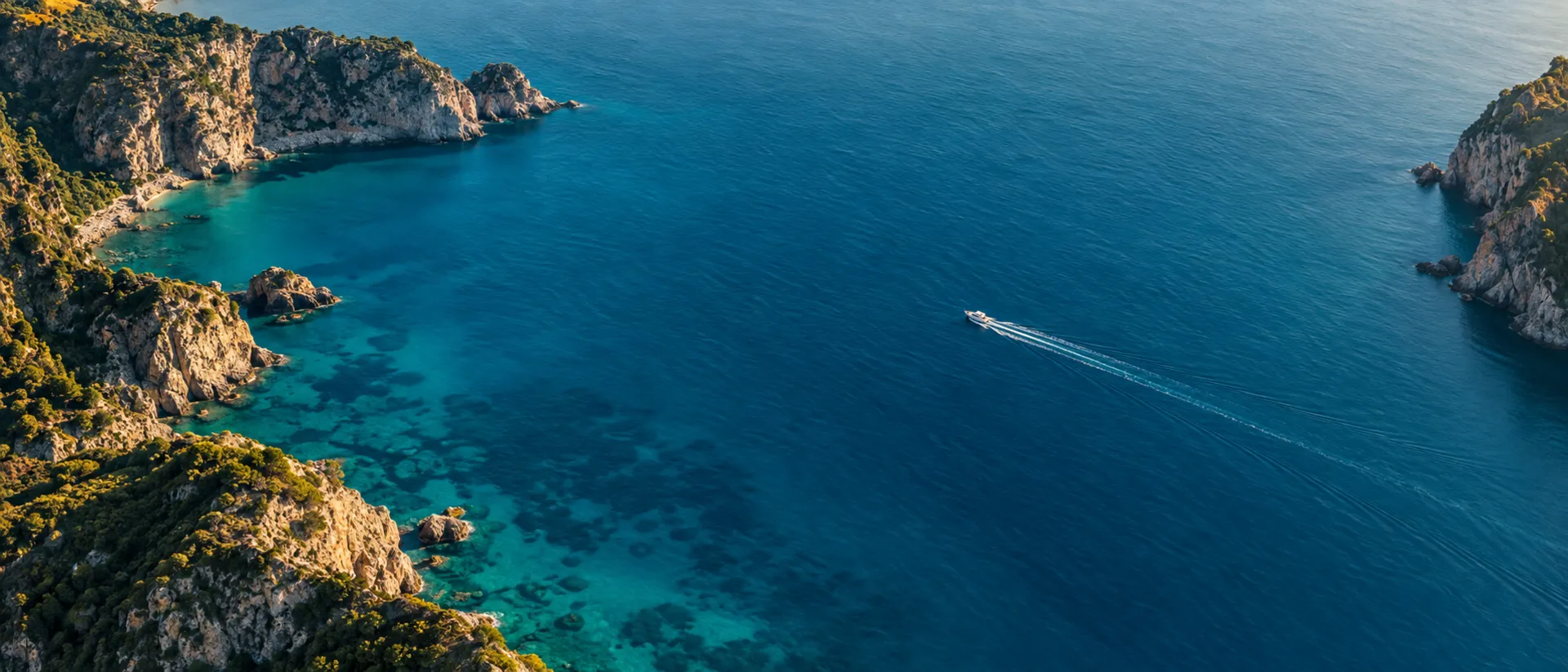 Aerial view of a deep turquoise Mediterranean bay with a single small boat crossing the water.